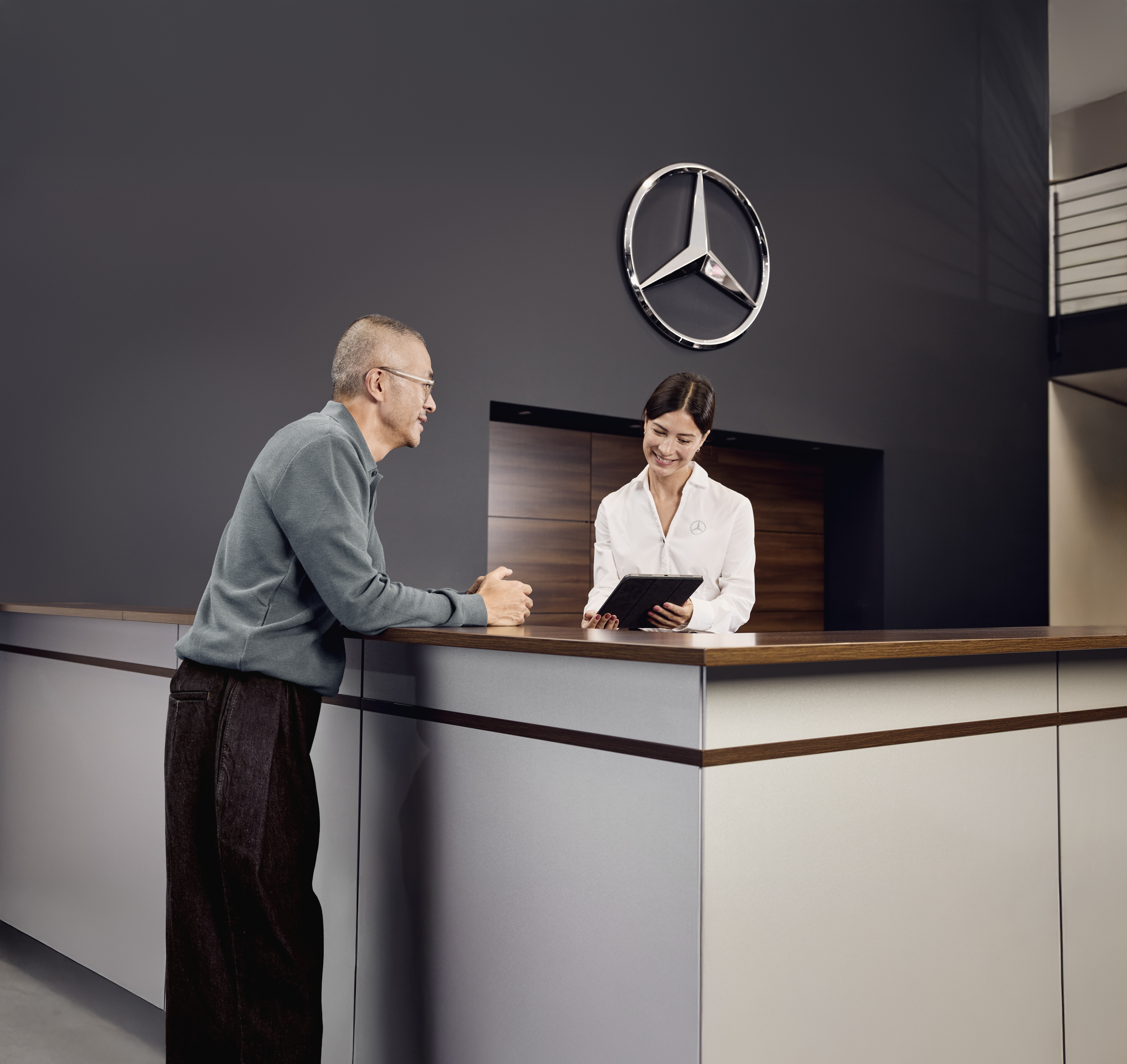 Consulta de Peças Genuínas | Mercedes-Benz Área de recepção de serviços em uma concessionária Mercedes-Benz, onde um cliente discute detalhes do serviço do veículo com um consultor de serviços na recepção.